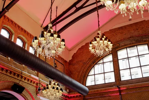 Mehrere Kronleuchter hängen an einer Backstein-Decke mit großen Fenstern und pinkem Licht.