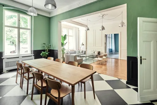 Großer heller Raum mit langem Holztisch, Stühlen und Schachbrettboden, angrenzendes Wohnzimmer.