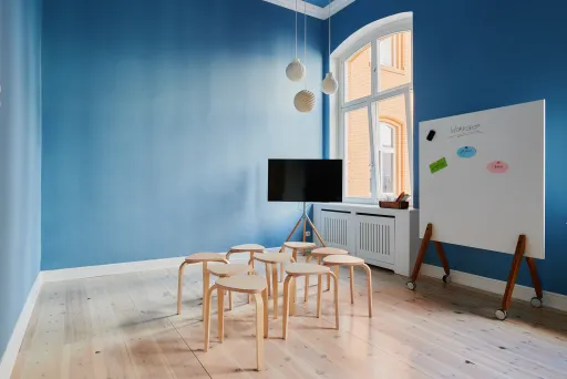 Heller Raum mit blauer Wand, Holzstühlen, TV, Whiteboard und großen Fenstern.
