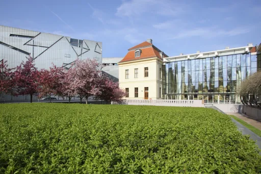 Moderne und historische Gebäude mit blühenden Bäumen und grüner Hecke im Vordergrund.