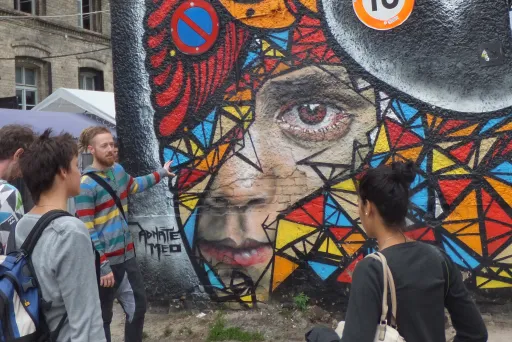 Bunte Graffiti-Wand mit Vogel- und Frauengesicht, mehrere Menschen davor.