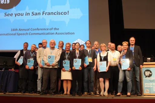 Gruppenfoto von Konferenzteilnehmern mit Urkunden auf einer Bühne.