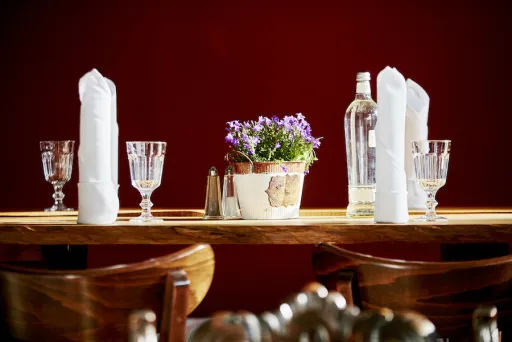 Holztisch mit Gläsern, Wasserflasche, Servietten und einem Blumentopf, roter Hintergrund.