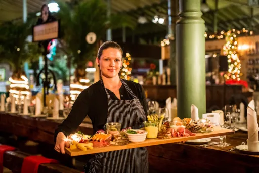 Kellnerin trägt ein großes Holzbrett mit verschiedenen Speisen in festlich dekoriertem Restaurant.