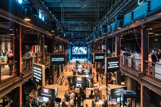 Große Messehalle mit Ständen, Besuchern und einer beleuchteten Bühne im Hintergrund.