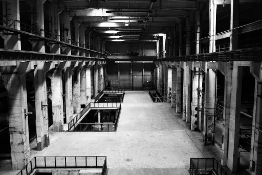 Große, leere Industriehalle mit Betonwänden und hohen Decken in Schwarzweiß.