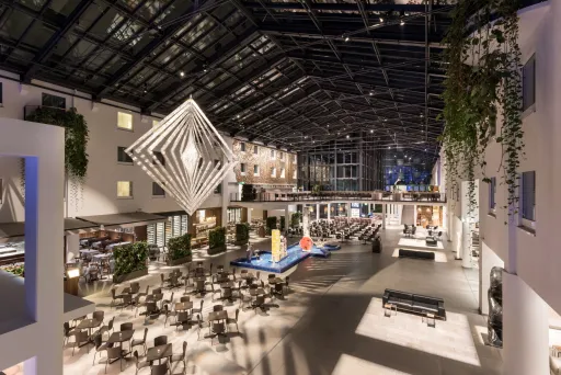 Große, moderne Hotel-Lobby mit Glasdach, vielen Tischen und geometrischer Skulptur.
