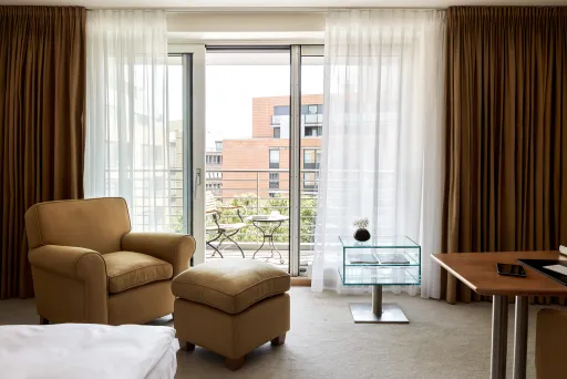 Helles Hotelzimmer mit Sessel, Ottomane und Balkonblick auf moderne Gebäude