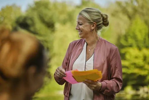 Frau mit rosa Jacke hält bunte Zettel und lächelt im Grünen.