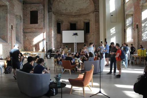 Menschen sitzen und stehen in einer hohen, lichtdurchfluteten Halle mit Ziegelwänden.