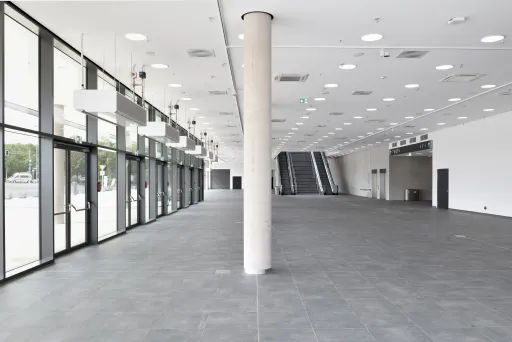 Heller, leerer Eingangsbereich mit Glasfront, Säule und Rolltreppen im Hintergrund.