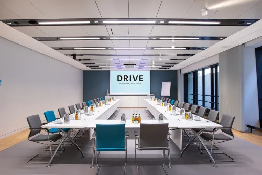 Moderner Konferenzraum mit U-förmigem Tisch, Stühlen und Bildschirm mit Schriftzug "DRIVE".