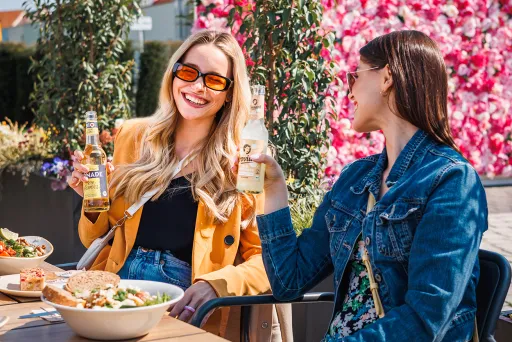Zwei Frauen stoßen lachend im Freien mit Getränken an, blühende Wand im Hintergrund.