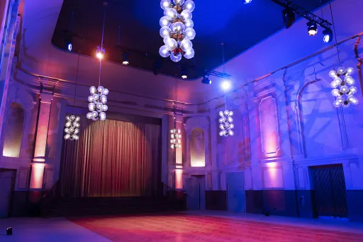 Der große Festsaal des Peter Edel mit Effektbeleuchtung, man sieht den Blick auf die Bühne, einen Bühnenvorhang sowie die neobarocken Zierelemente an den Wänden und die Deckenlampen, die im Stil des Palastes der Republik gehalten sind