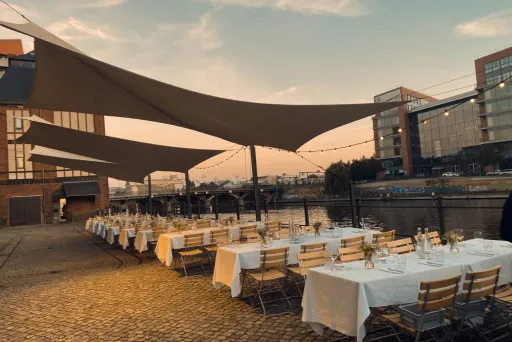 Lange, festlich gedeckte Tische draußen am Wasser bei Sonnenuntergang.