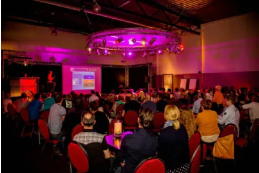 Viele Menschen sitzen im abgedunkelten Raum und sehen eine Präsentation auf Leinwand.