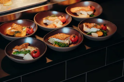 Sechs Teller mit Fischfilet, Gemüse und Kirschtomaten auf schwarzer Theke.