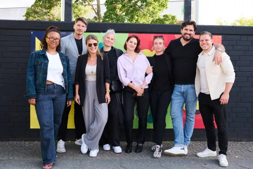 Acht Personen stehen lächelnd vor einer bunten Wand im Freien.