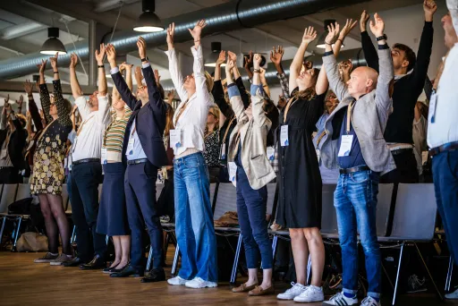 Viele Menschen stehen von ihren Stühlen auf und strecken ihre Arme in die Höhe.