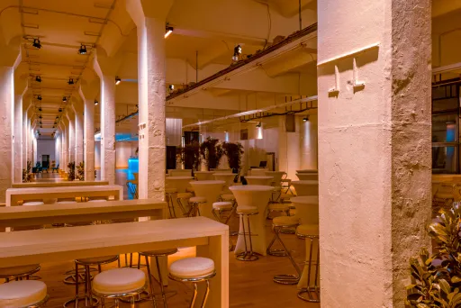 Modernes Restaurant mit hohen Decken, Holztischen und Barhockern, warm beleuchtet.