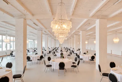 Großer, heller Festsaal mit runden Tischen, schwarzen Stühlen und Kronleuchtern.