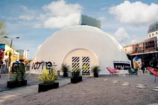 Weißes Kuppelzelt mit Pflanzen und bunten Stühlen auf einem Platz, blauem Himmel im Hintergrund.
