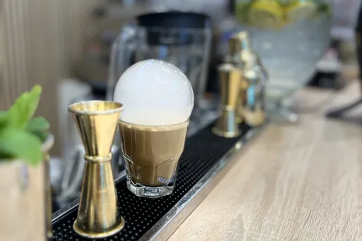 Latte mit Milchschaum auf Theke, daneben Messbecher und Glasgefäß mit Zitronenwasser.