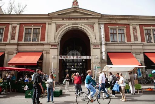 Menschen vor einem historischen Markthallengebäude mit roten Markisen und Ständen.