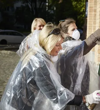 Women in plastic ponchos spray painting colorful graffiti on an outdoor canvas.