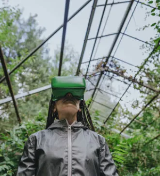 A woman with braided hair wears VR glasses in a greenhouse.