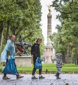 People walking in a park carrying blue trash bags, with trees and a monument in the background.