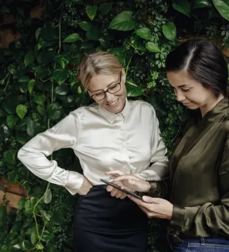 Two women in business attire stand in front of an ivy-covered wall and look at an Tablet with smiles on their faces.