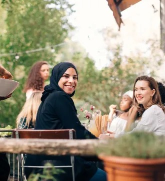 A group of young people are sitting outside a restaurant. A woman wearing a hijab smiles at the camera.