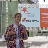 Eine Frau steht lächelnd und gestikulierend vor einer großen Bühne mit einem Banner, das „BESTIVAL“ sowie Veranstaltungsinformationen über das EUREF-Campus Berlin am 4. Juli 2025 zeigt. 