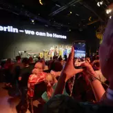 Teilnehmerin fotografiert mit dem Smartphone eine Bühnenperformance beim BESTIVAL 2025 in Berlin. Auf der Bühne steht eine Gruppe von Menschen, im Hintergrund leuchtet der Schriftzug „Berlin – we can be Heroes“. Das Publikum sitzt im abgedunkelten Raum.