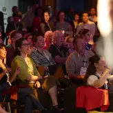 Publikum beim BESTIVAL 2025 in Berlin reagiert aufmerksam und begeistert auf einen Programmpunkt. Die Zuschauer:innen sitzen dicht beieinander, einige lachen oder gestikulieren – eine lebendige und mitreißende Stimmung im Saal.