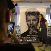 Eine Besucherin des BESTIVAL 2025 posiert vor einem Porträt von David Bowie mit dem Finger vor dem Mund. Die Szene wird von einer anderen Person mit dem Smartphone fotografiert. Im Hintergrund ist der Schriftzug #MEISTERSAAL zu sehen.
