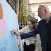 Ein Mann schreibt mit einem roten Marker auf eine große BESTIVAL-Wand, begleitet von einer lächelnden Frau. Die Szene spielt im Außenbereich der Alten Münze, geschmückt mit bunten Wimpelketten. Festivalstimmung und Austausch in entspannter Atmosphäre.