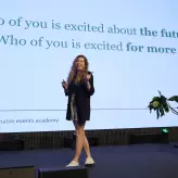 Rednerin der Sustainable Events Academy steht auf der Bühne des BESTIVAL 2025 und spricht vor großem Publikum. Hinter ihr ist die Präsentationsfolie mit der Frage „Who of you is excited about the future?“ zu sehen. Sie wirkt engagiert und motivierend.