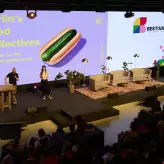 Bühnenpräsentation beim BESTIVAL 2025 zum Thema „Berlin's Food Collectives“. Zwei Speaker sprechen vor großem Publikum, im Hintergrund eine Leinwand mit Hotdog-Motiv.