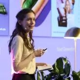 Eine Frau mit Headset-Mikrofon hält während einer Präsentation einen Vortrag. Sie steht auf einer Bühne mit violett beleuchtetem Hintergrund, auf dem Bilder und Texte zur Berlin Food Week zu sehen sind. In der Hand hält sie Notizen und einen Presenter.