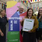 Vier Personen stehen nebeneinander vor einer bunten BESTIVAL-Bärenskulptur. Eine Frau hält stolz ein eingerahmtes Zertifikat (ISO 20121) in der Hand. Im Hintergrund sind bunte Wimpel, andere Besucher*innen und ein sonniger Innenhof zu sehen.
