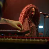 Eine Musikerin mit langen, lockigen Haaren steht auf einer dunklen Bühne vor zwei Mikrofonen. Im Vordergrund ist die Hand einer anderen Person an einem Keyboard zu sehen. Die Szene ist stimmungsvoll rot beleuchtet.
