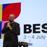 Staatssekretär Michael Biel auf der Bühne des BESTIVAL 2025 und spricht ins Mikrofon. Hinter ihm ist das farbenfrohe BESTIVAL-Logo sowie das Veranstaltungsdatum „3–4 July 2025“ auf der Leinwand zu sehen. Er Redner trägt dunkle Kleidung und Brille. 