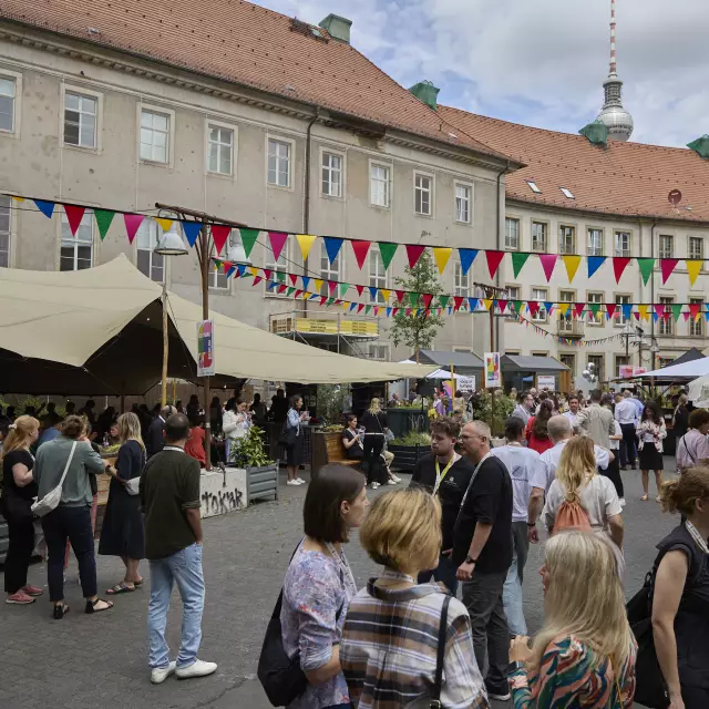 Buntes Treiben im Innenhof der Alten Münze in Berlin während des BESTIVAL 2025. Menschen schlendern zwischen Foodständen, Zelten und bunten Wimpelketten – im Hintergrund ist der Berliner Fernsehturm zu sehen. Sommerliche Festivalatmosphäre.