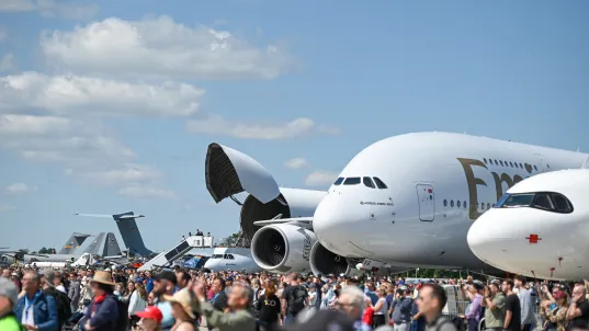 Large crowd at an airshow with airplanes, including an Emirates jet, on display under blue sky.