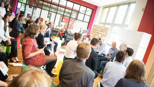 Menschen sitzen in einem hellen Raum und hören einer Diskussionsrunde zu.