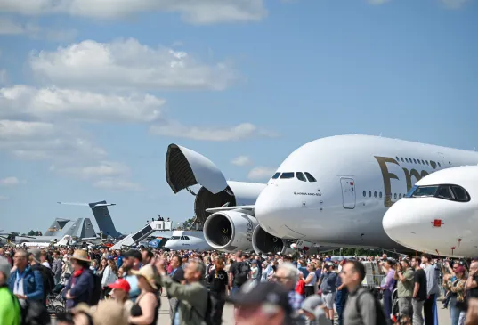 Large crowd at an airshow with airplanes, including an Emirates jet, on display under blue sky.