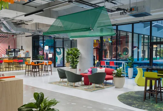 Modern office lounge with colorful furniture, plants, and large windows.