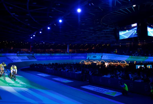 Cyclists on an indoor velodrome track under blue stage lights with a large audience watching.